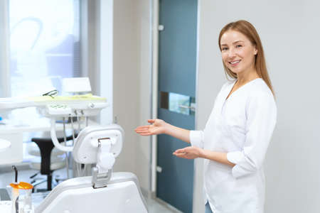 Portrait of smiling dentist standing in modern clinic office and inviting to sit in chair. Oral medicine concept. Stomatology chair and equipmentの写真素材