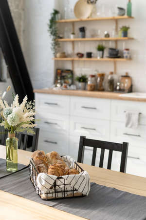 Fresh croissants in a basket and flowers in vase at wooden table in contemporary kitchen interior. Homemade bakery concept. Modern white furniture and decor on backgroundの写真素材