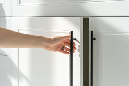Side view of woman's hand opening white wood cupboard door by black handle. Modern kitchen furniture...