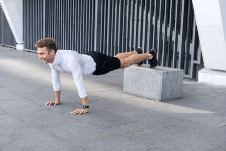 Side view of happy sportsman laughing, standing in plank pose, holding legs on box, hands on floor, making push ups exercise, training outdoors in the city. Wireless technology and sporty smart watchの写真素材