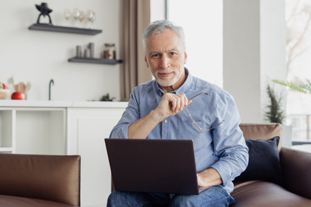 Portrait of elderly man with laptop sitting on couch. Mastering modern technologies for older...