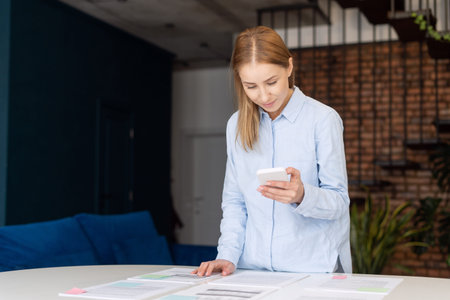 Portrait of beautiful woman solving business problems using smartphone. Variety of documents on...