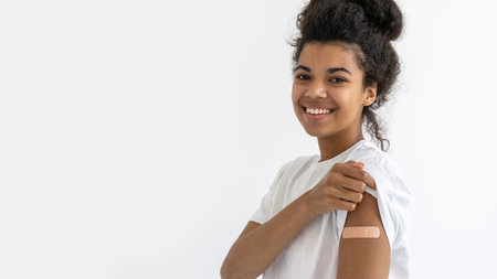 Cheerful african teenage girl having plaster on arm on white background. virus protection. Covid-19...