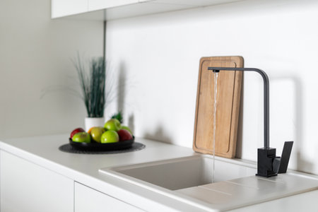 Side view of black sink with flowing water into Scandinavian kitchen interior design in white tones. Fresh fruits, cutting board and decor on background. home comfortの写真素材