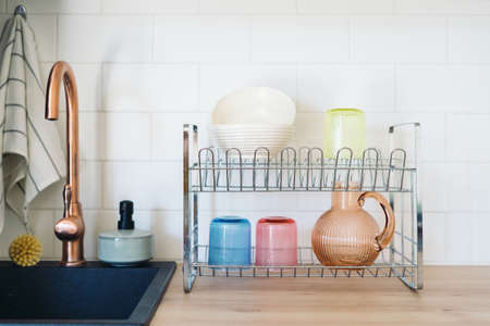 Metal drying rack for clean washed dishes with colored glass cups, jug for drinks and ceramic bowls, close to empty modern sink with copper faucet, dishwashing detergent soap, towel and wooden brushの写真素材