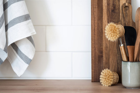 Close up view of ceramic cup with bamboo dishwashing brush, wooden spoon and cooking spatula on countertop. Kitchen utensils, tablecloth and cutting boards on shelf, against tiled wall with copy spaceの写真素材
