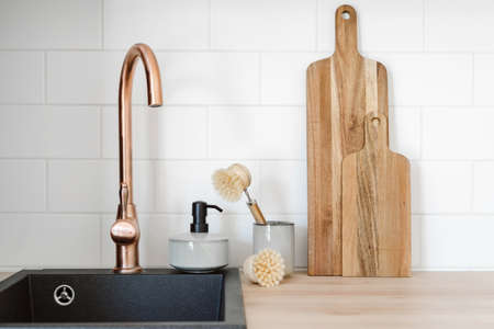 Wooden cutting boards, dishwashing brush in ceramic cup and soap detergent for dishes in dispenser bottle standing near black modern sink with copper faucet. Concept of household objects on kitchenの写真素材