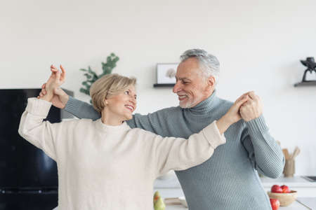 Happy and careless middle aged couple smiling and dancing in the kitchen together. Man looking at his gray haired wife with true love, holding her hands, enjoying weekend at homeの写真素材