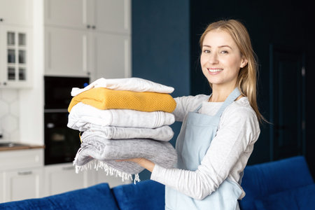 Entrust house cleaning chores to professionals. Portrait of smiling confident female folded clean and dry laundry clothes in stack, standing in room and look at cameraの写真素材