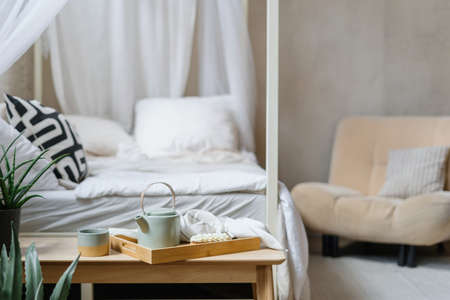 Selective focus on bamboo breakfast tray with teapot, cup of hot beverage and sweet eclair. Food and drinks on wooden bench against blurred cozy bed. Morning in hygge bedroom with boho chic styleの写真素材