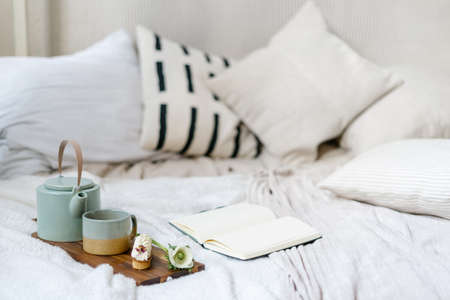 Open book or diary journal at comfort bed close to serving tray with breakfast. Notepad with blank pages near coffee cup, teapot and eclair on cutting board. Perfect morning in bohemian chic bedroomの写真素材