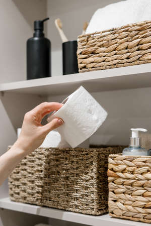 Cropped view of woman hand take toilet paper from wicker box. Domestic and household items on storage shelf in bathroom. Cosmetics bottles, clean and fresh towels in restroom closetの写真素材