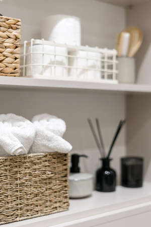 Organization of space in the bathroom cabinet. Selective focus on rolled fresh washed towels in wicker box in modern open shelf case. Toiletries in white closet or cupboard. Tidying up at home conceptの写真素材