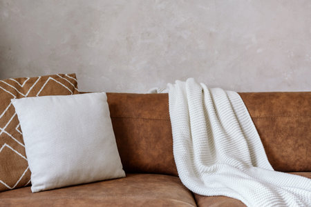 Closeup of brown eco leather couch with soft cushions and knitted white blanket near wall in room...