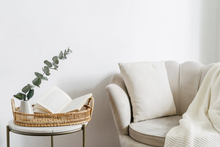 Wicker tray and open book close to eucalyptus branch in ceramic vase on side table. Bohemian design of living room with beige soft chair with cushion and white plaidの写真素材