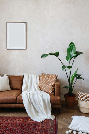 Vertical shot of cozy living room in bohemian apartment. Mock up poster hanging on wall over sofa with eco leather. Cushions and plaid on comfortable couch. House with potted plants and home decorの写真素材