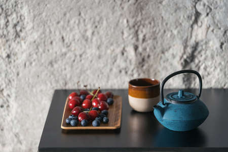 clay blue teapot with hot herbal beverage, ceramic cup of tea and wooden tray with fresh sweet cherry and blueberry on black countertop at kitchen roomの写真素材