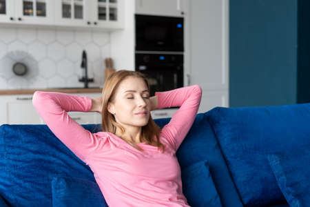 Peaceful and calm young woman with hands behind head relaxing on cozy couch in living room. Beautiful female with closed eyes leaning back, enjoying lazy weekend, sitting on sofa at home, taking napの写真素材