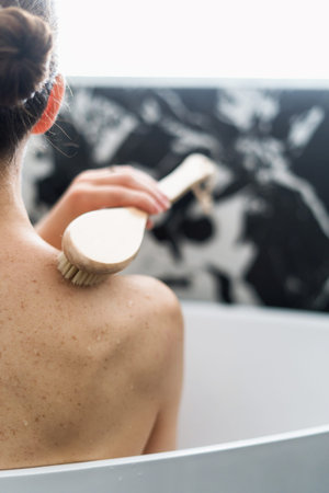 female naked back, hand with wooden massage brush for peeling body in bathtub in hotel bathroom or...