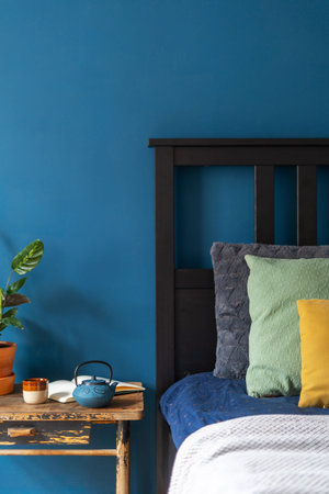 stylish interior design in lounge room with blue wall, comfort wooden bed with cushions and plaid, vintage side table, green potted houseplant, teapot and cup with herbal teaの写真素材