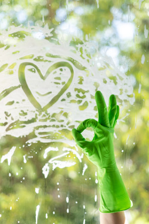 cropped shot of female hand in green protective glove showing okay gesture near window. heart symbol from soap foam. cleaning house with organic and eco friendly detergent. household service conceptの写真素材