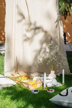 picnic in bohemian style at summertime on grass with pillows, flowers in vase, wicker tray with food and fruits. boho decoration with candles in garden, healthy breakfast on natureの写真素材