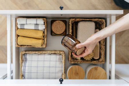 Top view of woman take recycled glass jar from open drawer in kitchen. Concept of organizing space at home. Storage box with towels, bamboo brush and cleaning sponge in wicker basketの写真素材