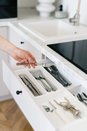Cropped view of woman hold metal fork in hand, standing on kitchen near white open cutlery drawer....