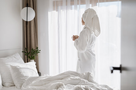Carefree woman with coffee cup in hands standing near window in hotel room. Female in white bathrobe enjoying weekend day in apartment, resting after beauty procedure. Advertising of resort and spaの写真素材
