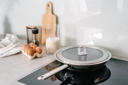 stainless steel pan with lid on glass ceramic cooker hob, eggs, milk, pepper and towel on countertop at kitchen with light interior, preparing breakfast at homeの写真素材