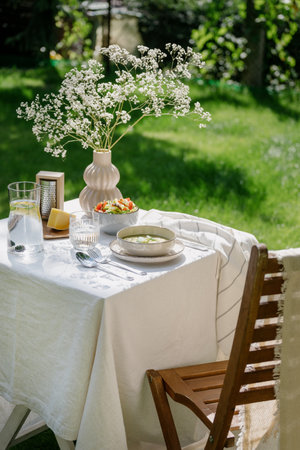 wooden chair and table serving white linen tablecloth, fresh vegetable salad, cheese, soup in bowl, cold water with lemon in glass jug and flowers in vase in park outdoors at summertimeの写真素材