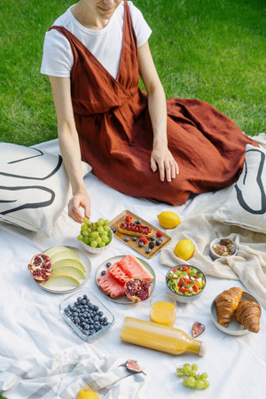 picnic in nature, female sitting on plaid serving with ripe melon, watermelon, blueberry, grape, sweet croissants, cake with berry and orange juice in bottle near glassful at sunny summer dayの写真素材
