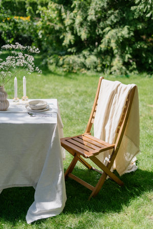 romantic breakfast in nature. wooden seat and table setting with white linen tablecloth, dinnerware, silverware, candles and flowers decor in vase in green park outdoorsの写真素材