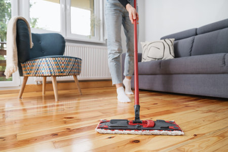 female hold red plastic mop in hands and washing parquet in room at modern apartment, cropped shot,...