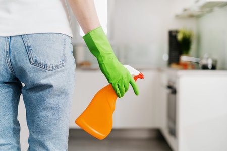female hand in green glove hold detergent spray in orange bottle with copy space at kitchen with...