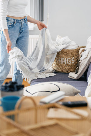 cropped shot of female in jeans take clean bed clothes or linen sheets from wicker basket in living...