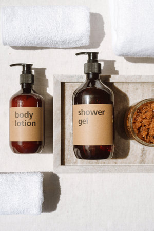 above view of shower gel and body lotion in brown plastic bottles with dispenser, exfoliating scrub in glass jar on marble tray and white folded terry towels at home bathroom, personal care conceptの写真素材