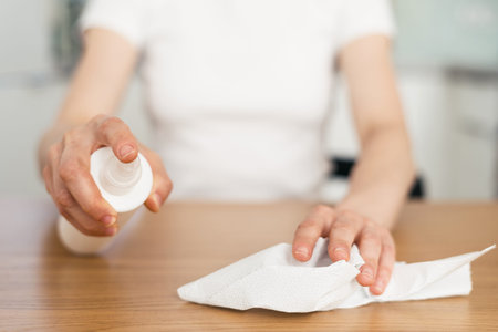 cropped shot of female hands hold detergent spray with tissue and wipe wooden table from dust in...
