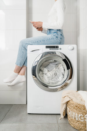 female housewife sitting with mobile phone on automatic washing machine with bed linen inside and closed door, wicker basket with clothes on floor in bathroom, cropped shotの写真素材