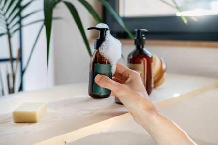 Close up shot of woman hand taking brown recycled glass bottle with soap dispenser from wooden shelf. Caddy tray with cosmetics products over bath. Skin care and morning routine conceptsの写真素材