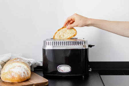 electric toaster on black countertop at home kitchen with white wall and female hand put slice bread for making crispy toast for breakfast, cooking conceptの写真素材