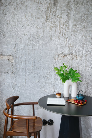 ceramic vase with leaf branch, sweet cherry on plate, teapot with cup and diary on round table and brown wood chair at stylish apartment with loft interiorの写真素材