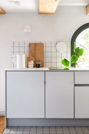 modern cabinet, induction hob, electric kettle and cups on countertop, white tile on wall and houseplant near window in kitchen with stylish interiorの写真素材