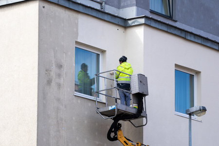 Maintenance worker in workwear using roller brush paints the facade of a residential house. Man in overalls standing outside building and repainting concrete wall close to window. Improvement conceptsの写真素材