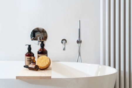 massage brush, organic skincare cosmetics, brown dispenser bottles with body lotion and shower gel on bamboo caddy on bath tub in luxury apartmentの写真素材