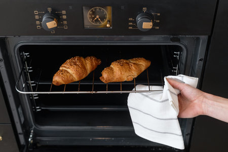 cropped shot of female hand holding warm fresh croissants with crust on metal oven rack at modern...