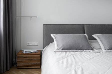 soft pillows and cotton blanket on bed with gray headboard, open book on wooden side table and metal lamp in room with modern interiorの写真素材