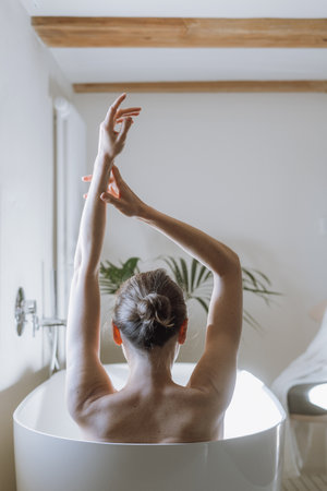 Back rear view of young woman taking a bath, raising hands up, enjoying morning in hotel bathroom. beauty concepts. Skincare, bodycare and self care routineの写真素材