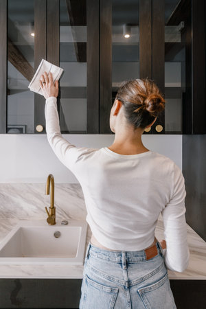 back view of housewife with cloth in hand cleaning wooden door of cabinet at kitchen with dark...