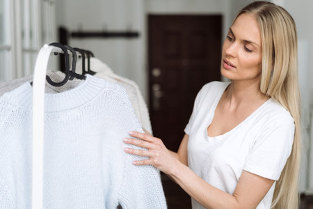 Concept of basic wear and capsule wardrobe for winter season. Young woman standing near open wardrobe with clothes, choosing wool knitted sweater in pastel colours. Female getting dressed at morningの写真素材